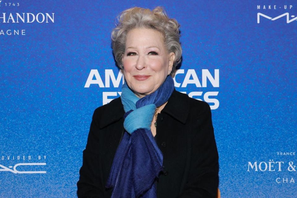 Bette Midler poses in front of a branded backdrop wearing a dark blazer and a layered scarf around her neck