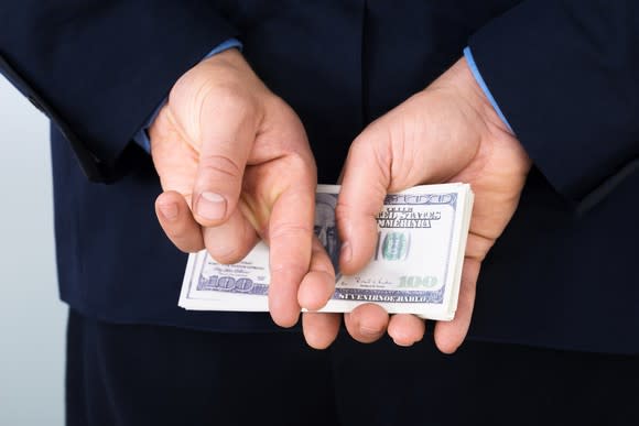 A businessman in a suit hiding a stack of hundred dollar bills behind his back, with his fingers also crossed.