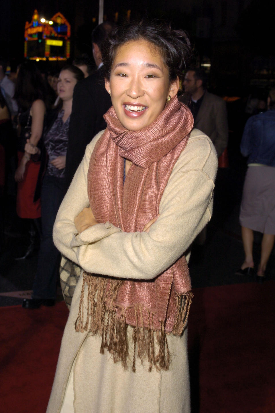 Sandra Oh smiling on a red carpet, wearing a long coat and a scarf