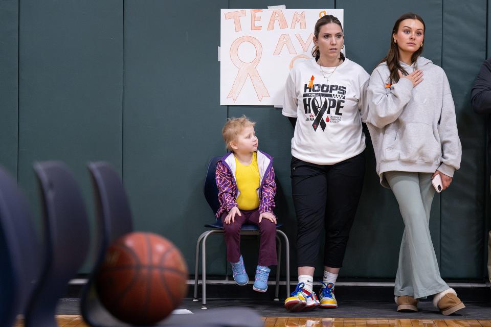 Ava Blazis, a 5-year-old Sutton native with acute lymphoblastic leukemia, sits during the national anthem as her aunt, Johanna Annunziata, and mentor Lily Sullivan stand. Sullivan, now cancer free, had the same diagnosis as Ava.