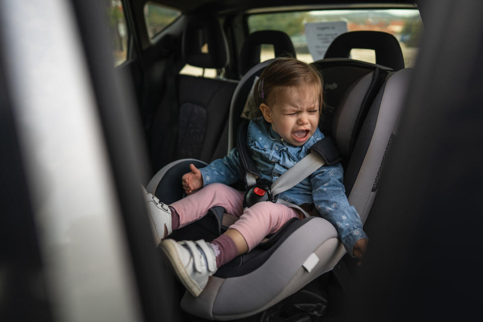 A toddler sits crying in a car seat, wearing a denim jacket and pink pants. The child appears upset and holding a partially eaten snack in one hand