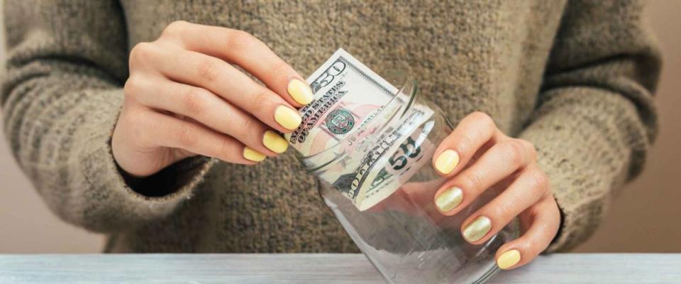Girl in brown sweater with yellow manicure puts money in a glass jar, close-up