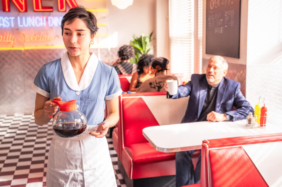 Waitress in a 1950s-style diner holds a pot of coffee. A man in a booth holds a mug. Other diners are in the background