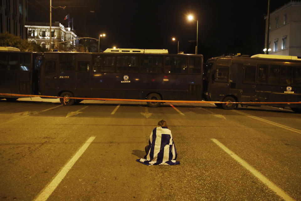 An opponent of Prespa Agreement, draped in a Greek flag, sits on a road blocked by riot police buses, following a rally outside the Greek Parliament, in Athens, Thursday, Jan. 24, 2019. Greek lawmakers are debating a historic agreement aimed at normalising relations with Macedonia in a stormy parliamentary session scheduled to culminate in a Friday vote. (AP Photo/Thanassis Stavrakis)