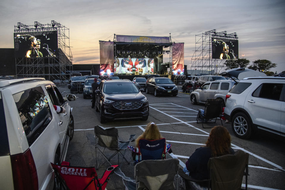 FILE - Attendees sit next to their car as J.R. Moore & Zack Mack perform at the Drive-In at Westland Mall in Columbus, Ohio on Oct. 10, 2020. After a historic season, winter is coming at the drive-in. Summer and early fall have seen the old drive-in transformed into a surprisingly elastic omnibus of pandemic-era gathering. It has hosted concerts and comedy shows, business conferences and Sunday services, graduations and weddings. Drive-ins in Texas, California and Florida can keep humming all year but most of the U.S.’s roughly 300 drive-ins are seasonal. They aren’t built for the cold, and they’re definitely not built for the snow. (Amy Harris/Invision/AP, File)