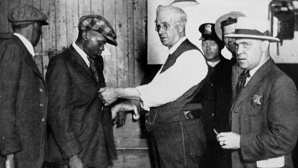 PHOTO: Officers search a Black man for arms at a police station in Chicago, Illinois, in 1922. (New York Public Library)