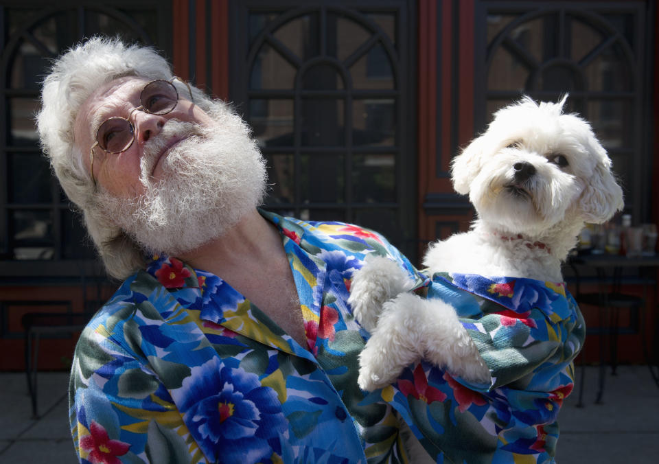 Elderly man with a bushy white beard, wearing circular glasses and a floral-patterned shirt, holds a white fluffy dog dressed in a matching shirt