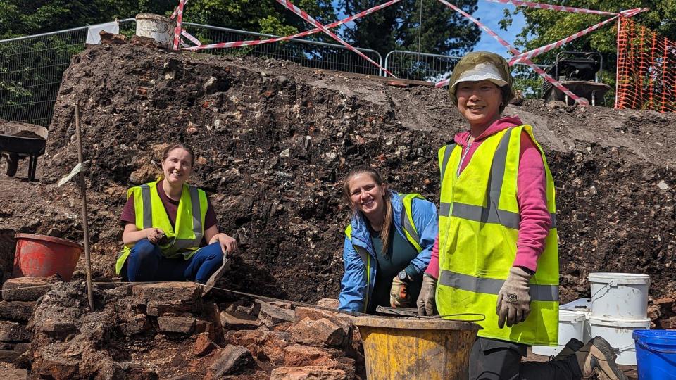 Three women smiling to the camera in the middle of an archaeological dig