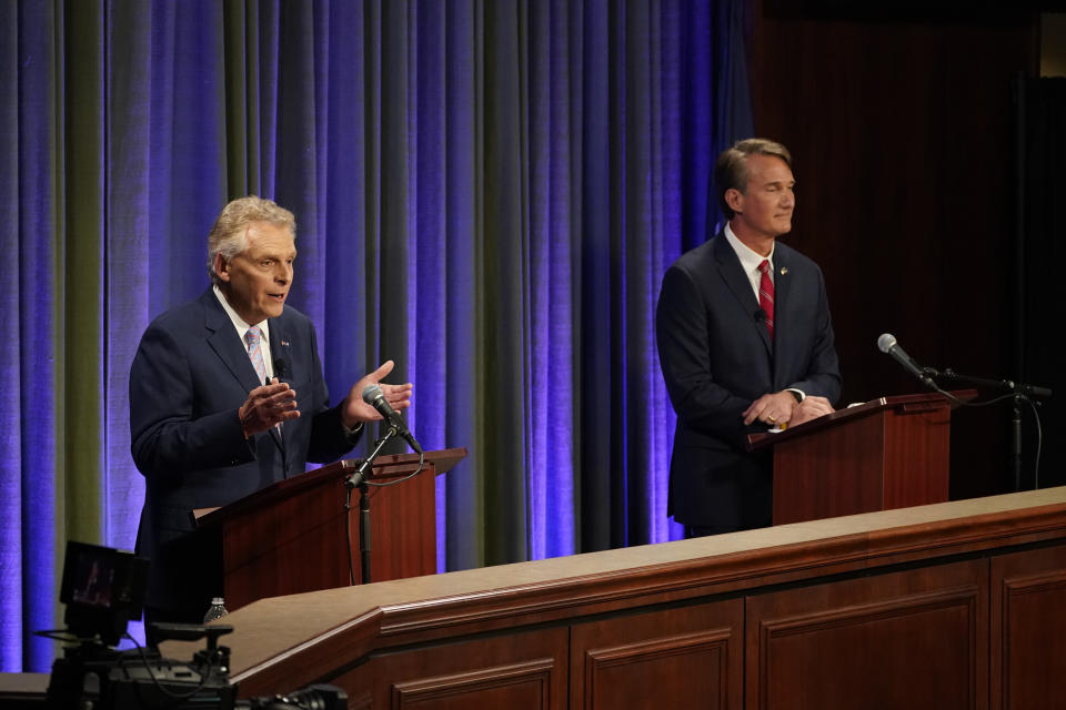 Terry McAuliffe and Glenn Youngkin stand at wooden podiums with microphones in front of a purple curtain at a debate.