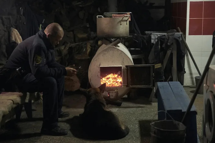 An emergency worker and his dog warm up in front of a wood-burning oven in a shelter in Bakhmut, the site of the heaviest battles with the Russian troops, in the Donetsk region, Ukraine, Friday, Dec. 9, 2022. (AP Photo/LIBKOS)