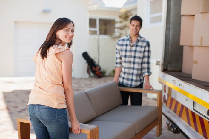 A young woman and young man, who are carrying a sofa, standing next to an open-backed van with a house in background.