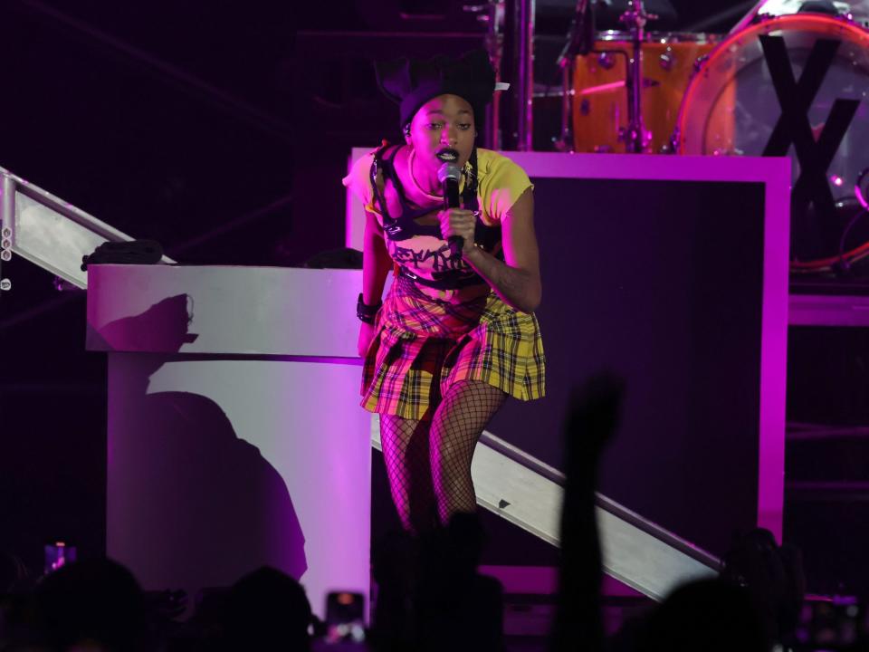 Willow is onstage swearing a yellow baby tee, a black leather harness over her tee, yellow plaid skirt, fishnet stockings. She also has black lipstick and a black beanie on, She's leaning towards the crowd, singing into the mic in her hand.