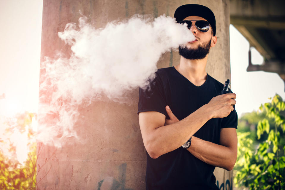 A young man with a beard and sunglasses blowing vape smoke out of his mouth while outside.
