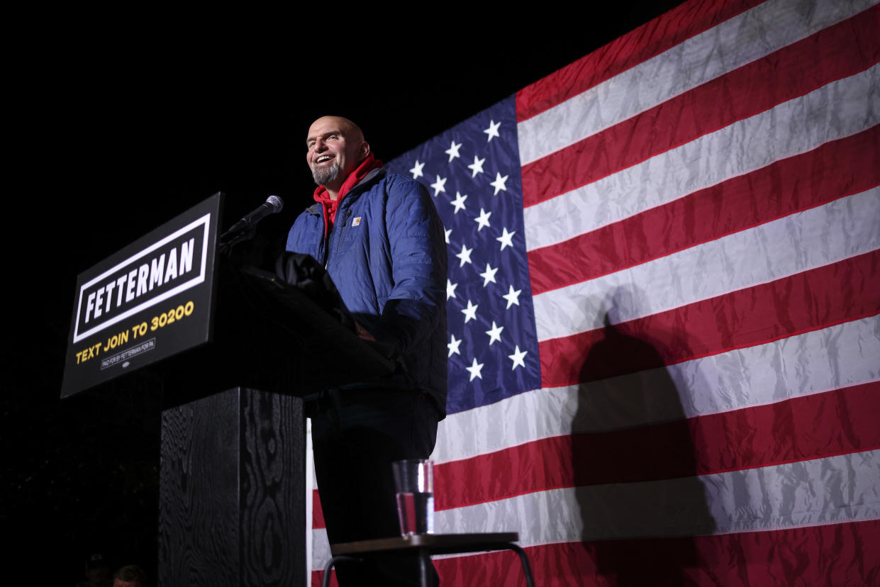 U.S. Senate candidate John Fetterman onstage at a campaign event with an American flag in the background.