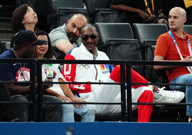 Snoop Dogg, in a red, white and blue tracksuit and wearing sunglasses. sits with his legs crossed in the crowd at the gymnastics. 