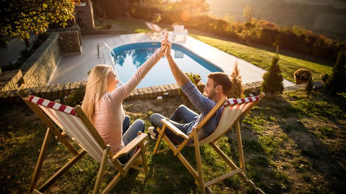 Young happy couple relaxing in deck chairs at their back yard and holding their arms together.