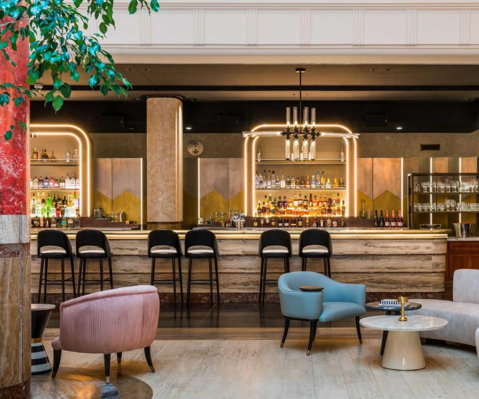 Inside Kimpton Margot Sydney's art deco bar with dark bar stools sitting under the pale bar, chandeliers above and coloured velvet arched lounges and chairs in the foreground.