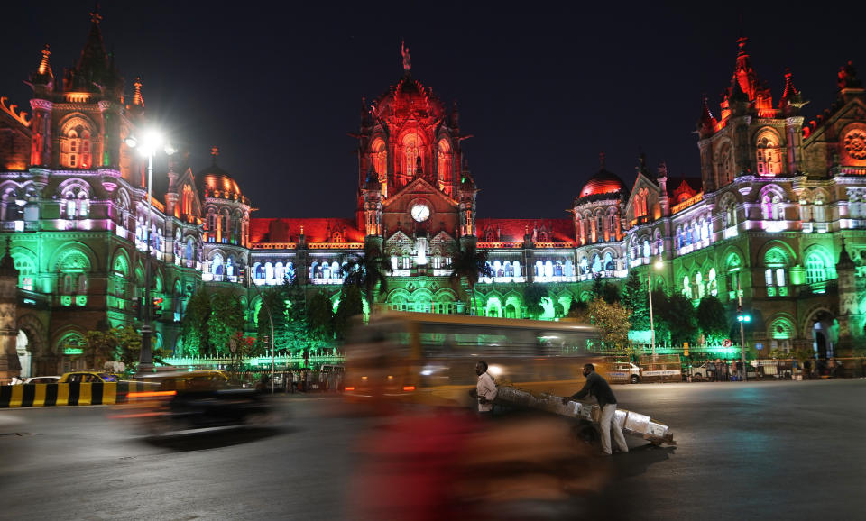 Two men transport goods on a pushcart as vehicles move past the Chhatrapati Shivaji Maharaj train terminus building that is illuminated on the eve of India's Republic Day in Mumbai, India, Tuesday, Jan. 25, 2022. (AP Photo/Rajanish Kakade)