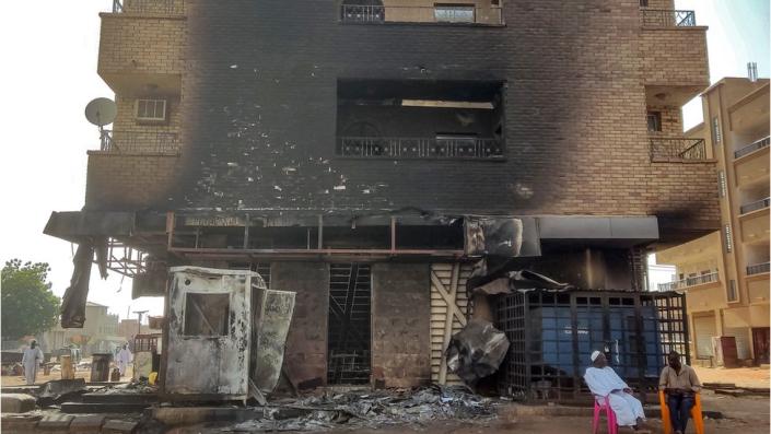 Building charred from and attack, The ground floor is completely destroyed. Two men sit on colorful bright chairs at the bottom of the building.