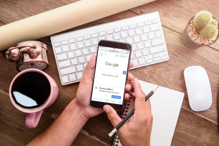 Overhead view of a person's hands holding a cell phone with Google's home page showing. Various desk-top items, such as a keyboard, shown.