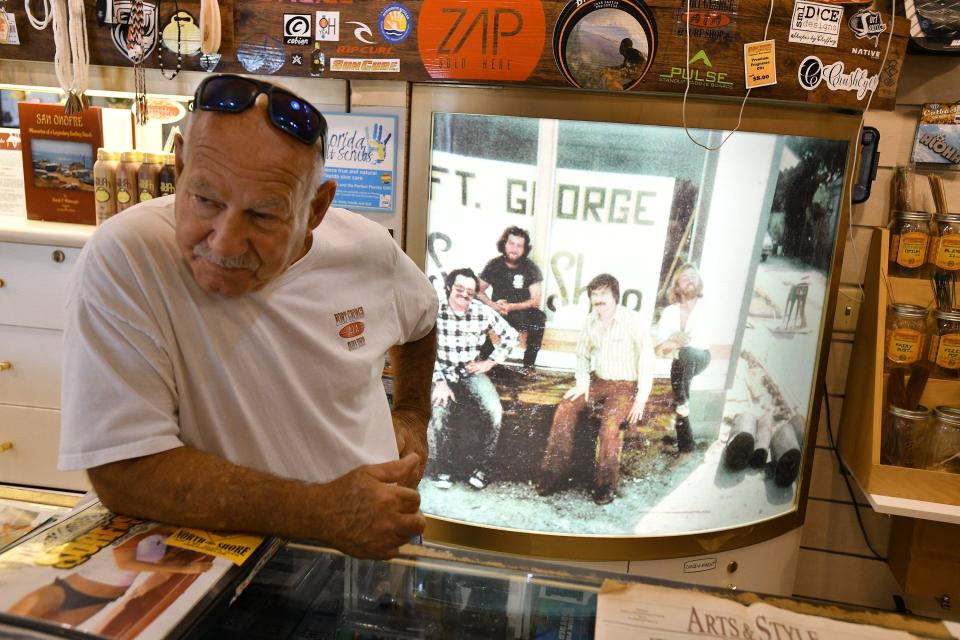 Jim Rodgers talks with a guest at the front counter of his Fort George Surf Shop. The backlit photograph behind him is a photo of himself and friends at the shop in the 1970s. Rodgers is the one in the black T-shirt.