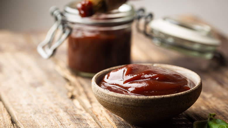 Barbecue sauce in dish on wooden table next to glass jar and brush