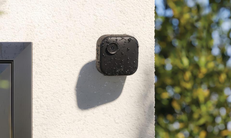 Amazon marketing photo of the Blink Outdoor 4 camera. The black camera has a flat face and rounded corners. It's mounted on a light-colored stucco (outdoor) wall with blurred trees in the background.