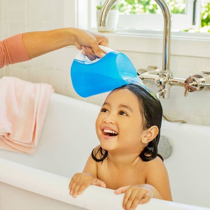 Adult pouring water from blue pitcher over smiling child's head in a bathtub, demonstrating hair washing