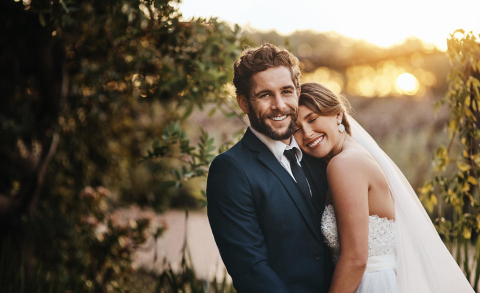 A couple, an unidentified man in a suit and an unidentified woman in a wedding dress, are smiling and embracing outdoors with trees in the background