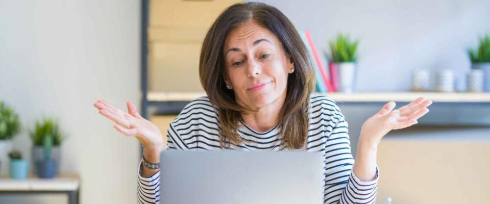 Middle age senior woman sitting at the table at home working using computer laptop clueless and confused expression with arms and hands raised. Doubt concept.