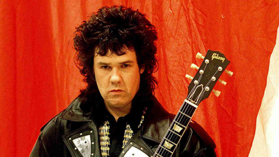 Gary Moore holding a guitar against a red background in 1988