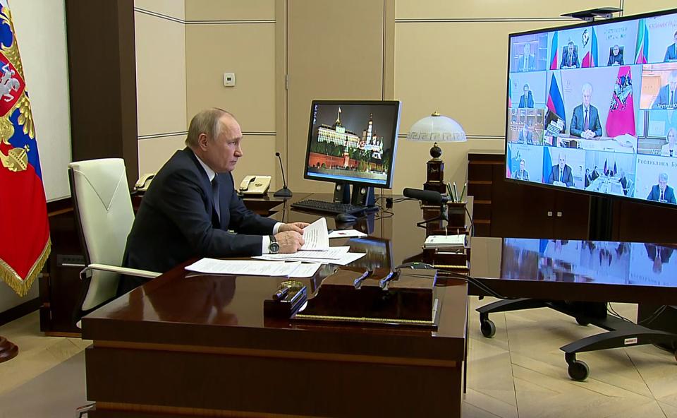 Putin faces a large monitor with the images of numerous people at a video meeting.