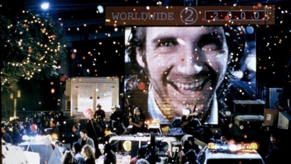 Ralph Fiennes' face appears on a giant video screen amid a New Year's Eve celebration in Time Square, with dozens of people and cars littering the street and Christmas lights on buildings, in one of the best virtual reality movies, Strange Days.