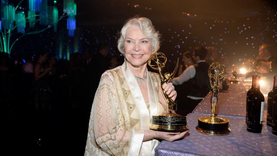 ellen burstyn smiles and looks to the left of the camera, she holds her emmy award while standing next to a table with a purple tablecloth, she wears a cream, tan and gold caftan