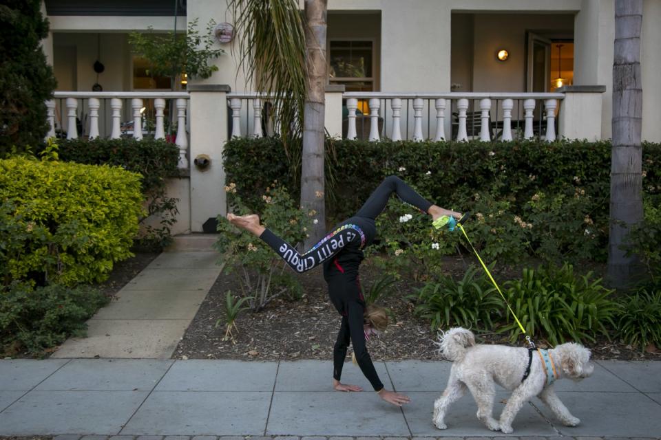 A girl walks on her hands along the sidewalk, with her dog's leash held by her toes.