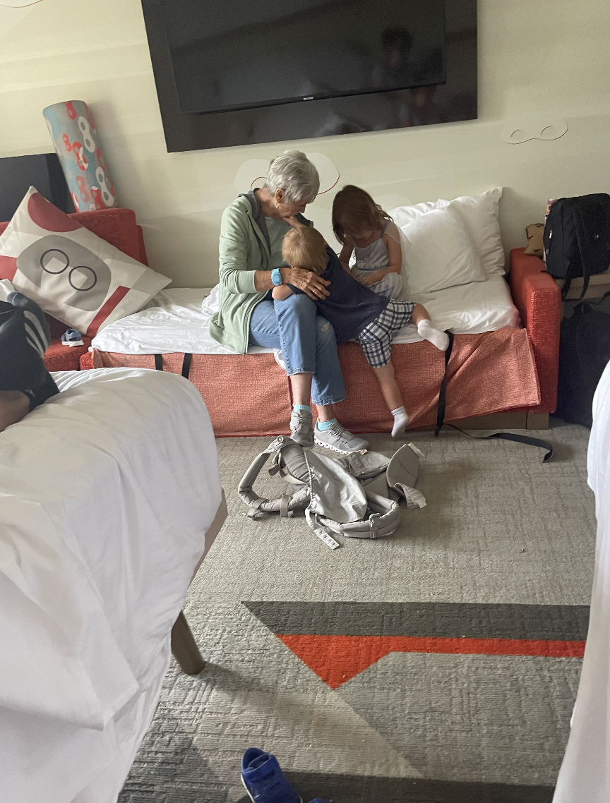 An older woman and two children sit on a couch in a room. They are engaged in a quiet, intimate moment. A backpack lies on the floor in front of them