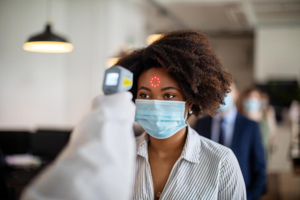 A technician in a white coat holds up scanner to project a circle of points of light onto the forehead of a Black woman wearing a surgical mask, as other people in masks line up behind her in a large room in an office with overhead lights.