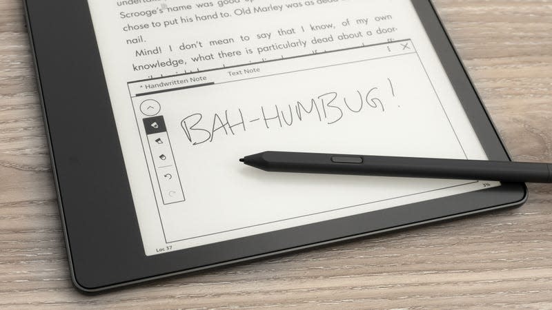 A sticky note added to an ebook on the Kindle Scribe's screen with a hand-written "BAH-HUMBUG!" message, with the device sitting on a wooden table.