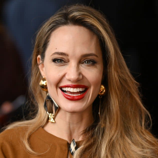 Angelina Jolie, smiling and wearing gold earrings and a brown outfit, attends an event