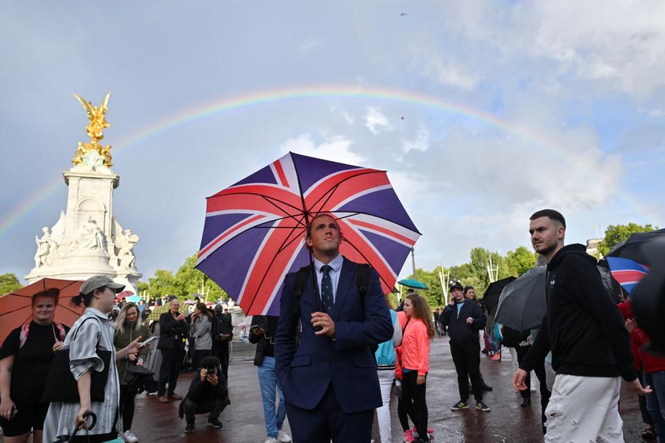 A man looks on holding a Union Jack umbrella as a rainbow is seen outside of Buckingham Palace in London.