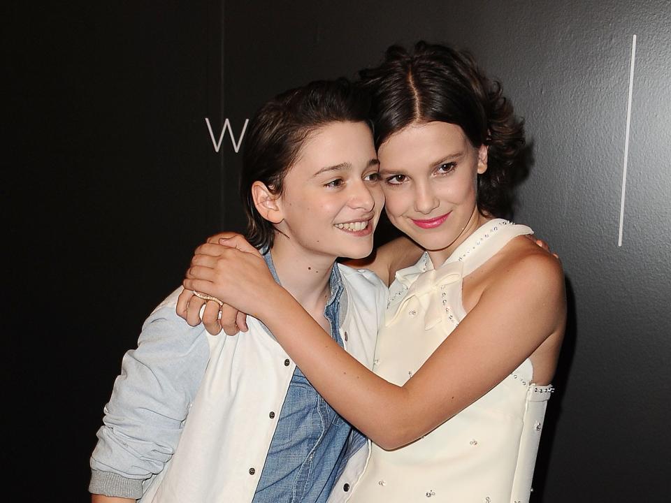 a young millie bobby brown and noah schnapp posing together on a red carpet, their arms around one another as they smile towards a camera.