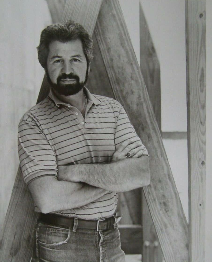 Black and white image of Bob Vila with full beard, wearing jeans and a striped shirt.