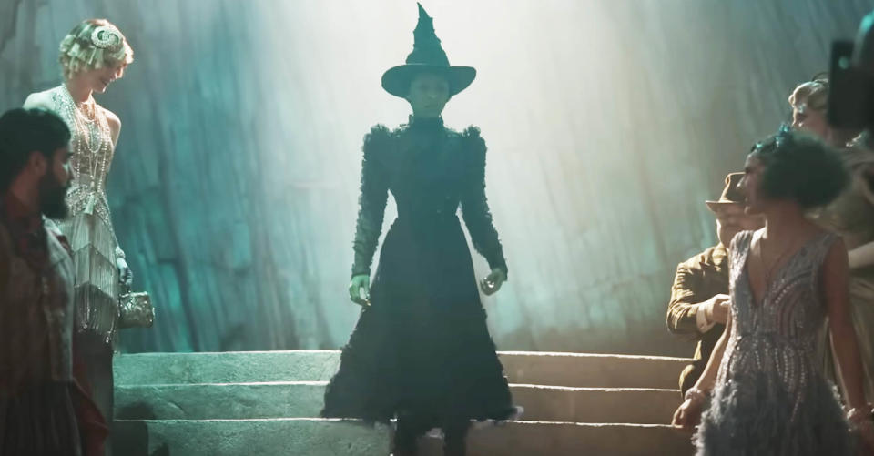 Elphaba coming down the stairs at a party wearing a long dress and pointy witches hat