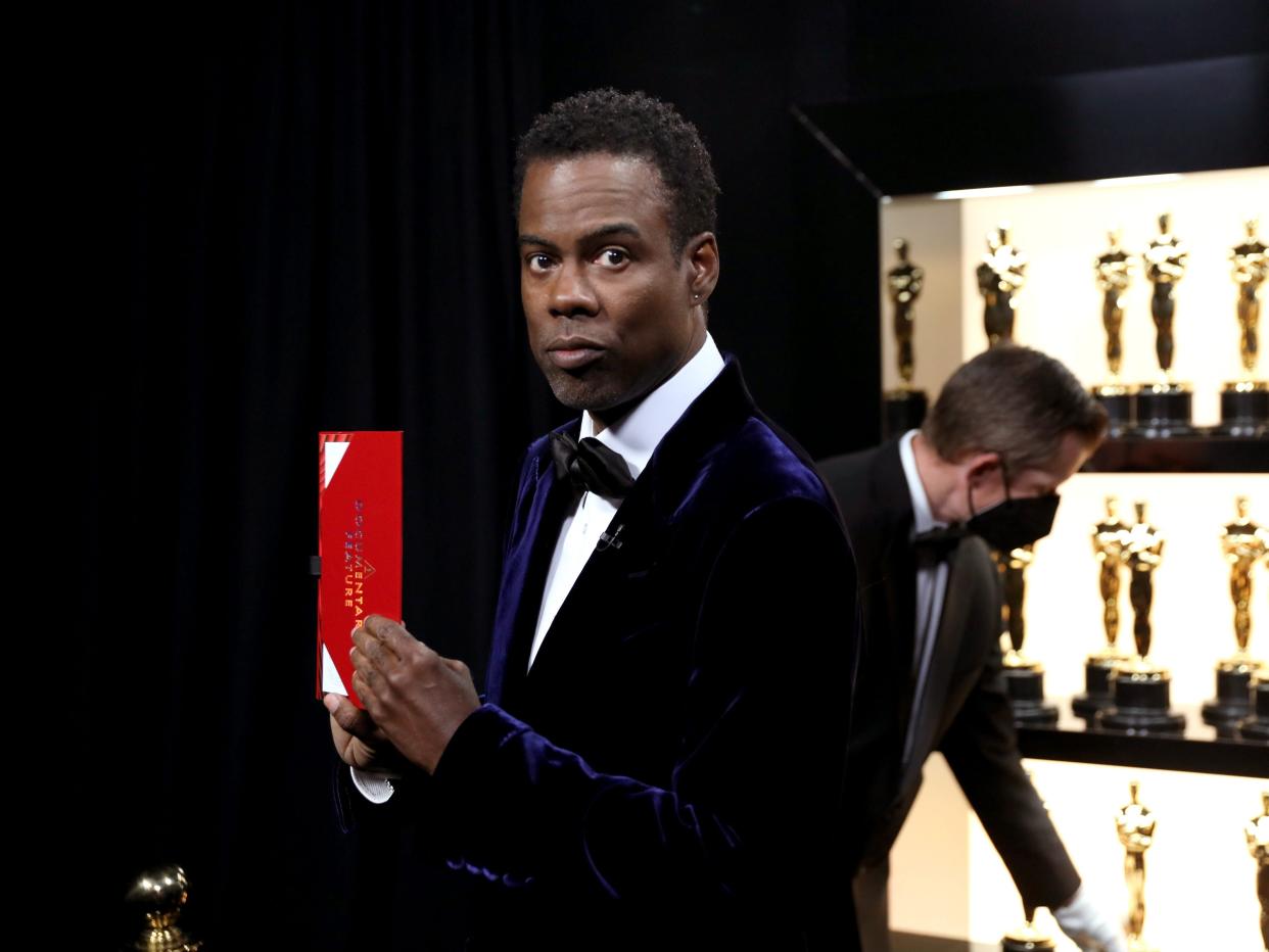 Chris Rock holding an Oscar winner envelope with Oscar statues behind him