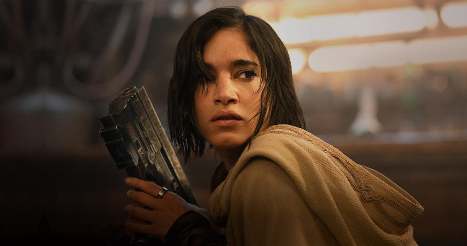a young woman in a cloak holds a futuristic pistol