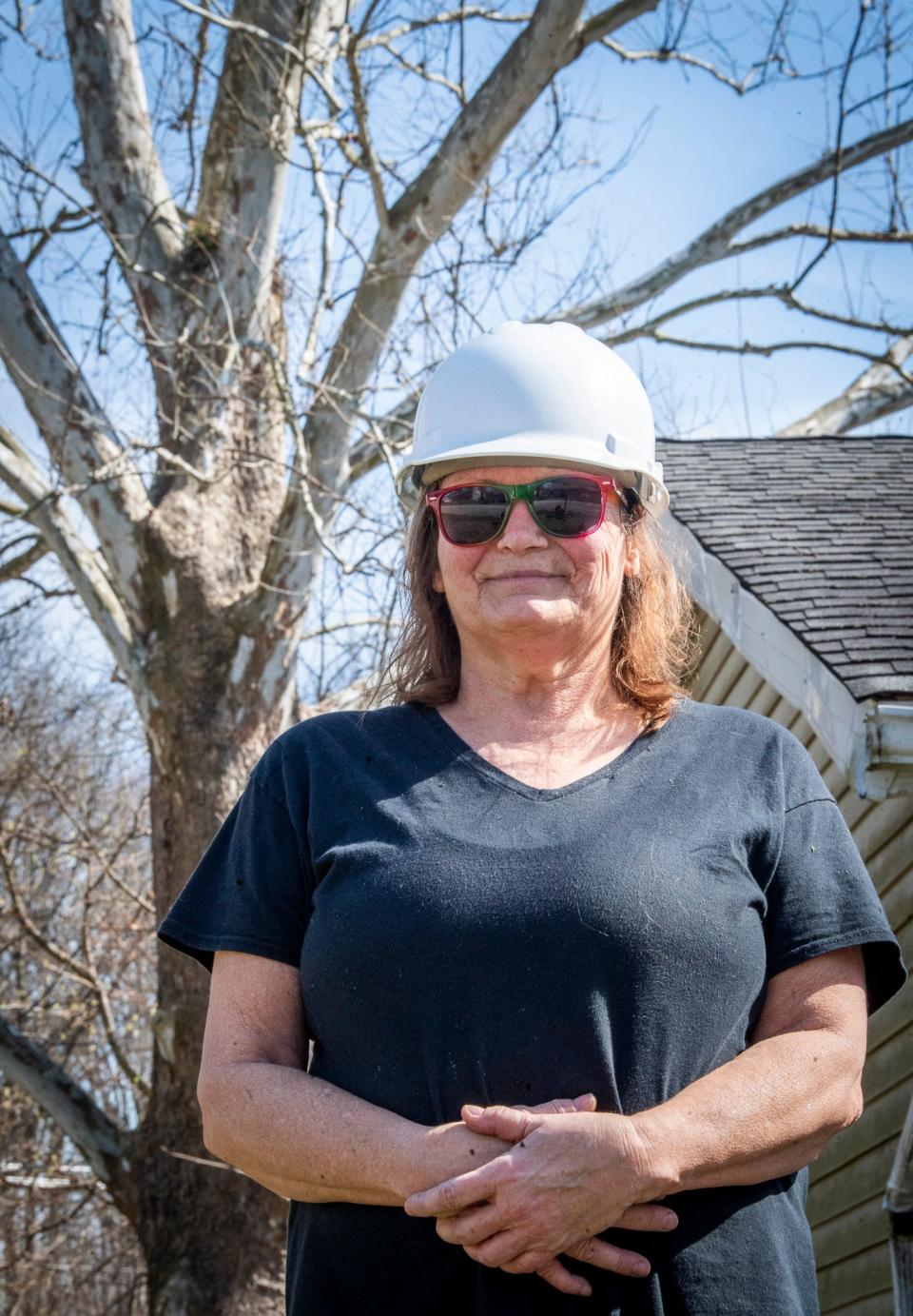 Angela Conner stands in her yard wearing a hard hat that helps protect her from nesting hawks at her house in Ellettsville on Tuesday, March 12, 2024. Conner has been repeatedly attacked by a red-shouldered hawk that lives in the sycamore tree in her backyard, and can be seen behind her. The nest, though small, is in the top "Y" split of the tree behind her.
