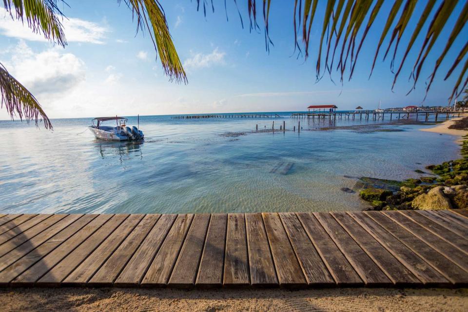 A scenic beach view showing a wooden boardwalk at the waters edge, framed by palm leaves. The crystal clear water against a bright blue sky.