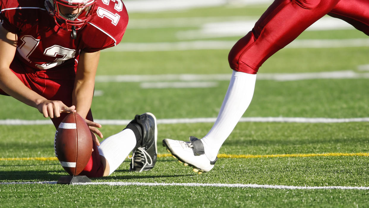 A football player tees up the ball for the approaching kicker, both on red jerseys