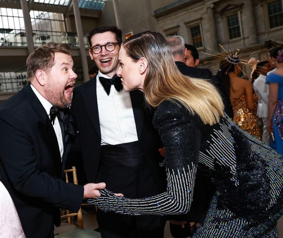 Three people at an event, one male in tuxedo and glasses looking amused, and a male and female in formal attire engaging excitedly