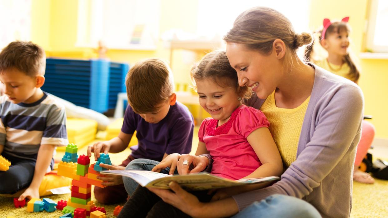 Happy little girl sitting in teacher's lap at preshool and reading a book while other children are playing.